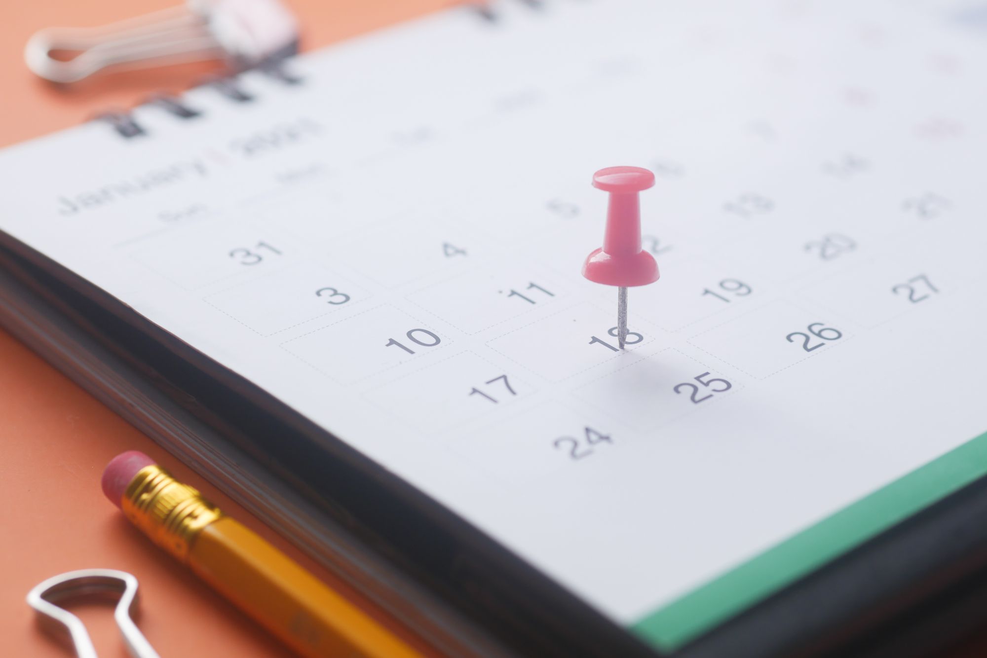 Gros plan sur une page de calendrier avec une punaise rose, un crayon et une pince à dessin sur un fond orange.